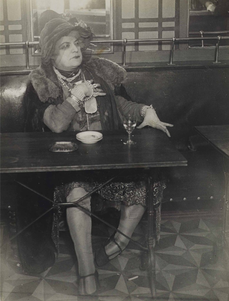 La Môme Bijou with her hand resting on her chest. She looks away from the camera as she sits with a drink.