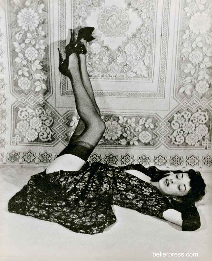 Holly Faram photographed by John Willie. She is laying down, and her dress is riding up. She is lifting her black stockinged legs, and wears extremely high black leather heels and black leather opera gloves.