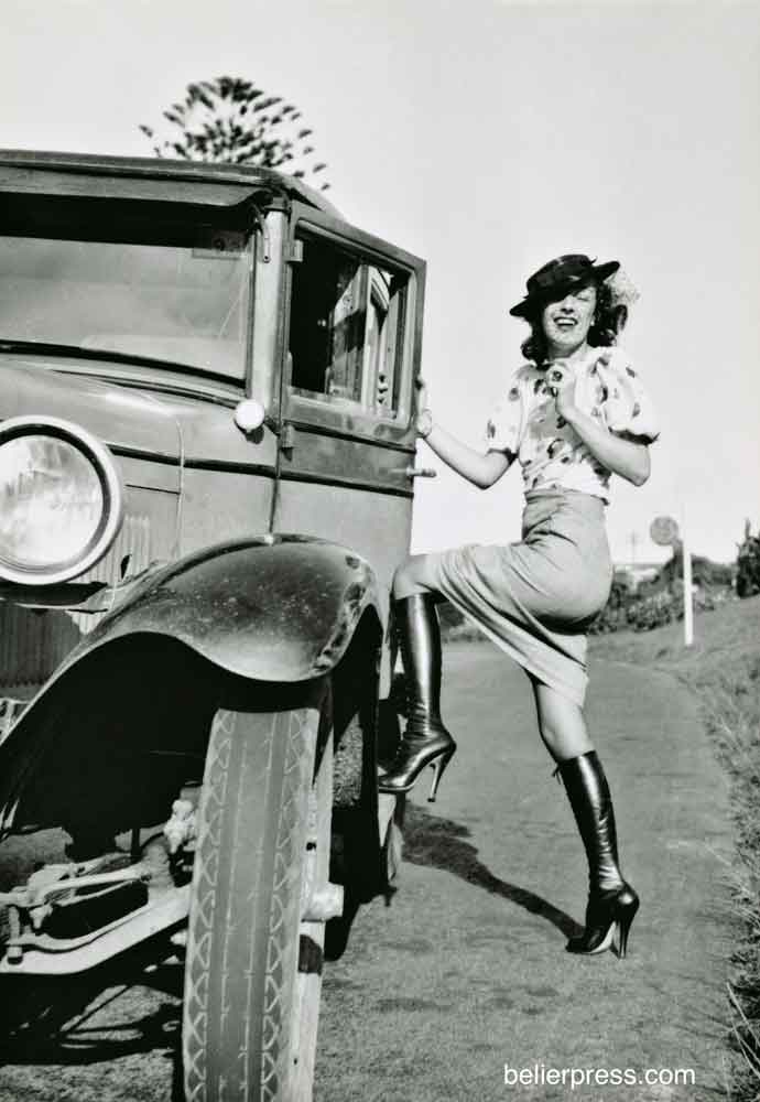 Holly Faram photographed by John Willie. She is stepping into an automobile and laughing. She wears a tight pencil skirt and knee high, high heeled leather boots.