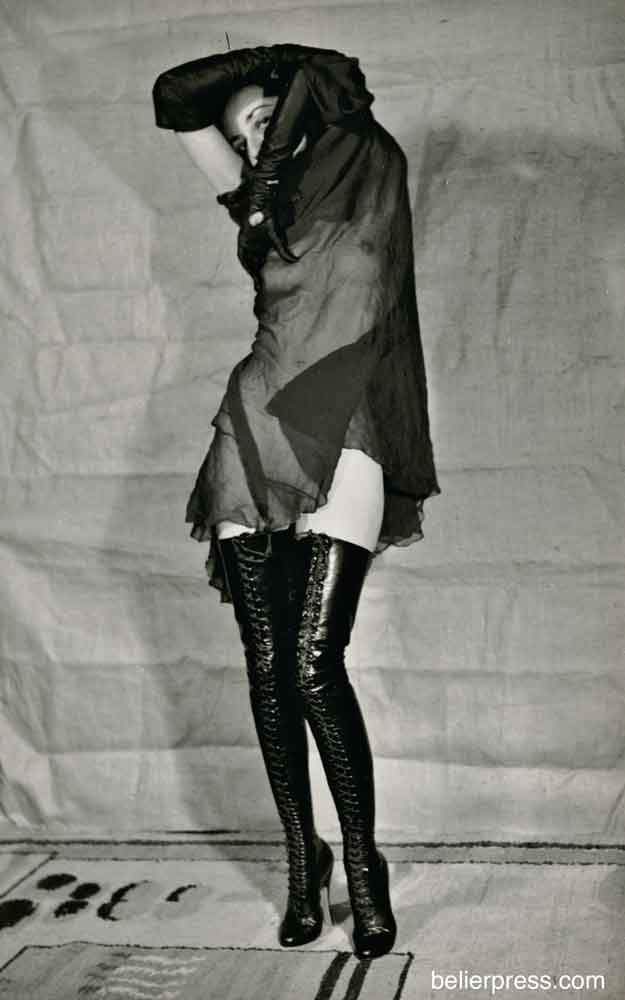 Holly Faram photographed by John Willie. She holds sheer black fabric over her nude body. She is wearing thigh high black leather boots and long black opera gloves.