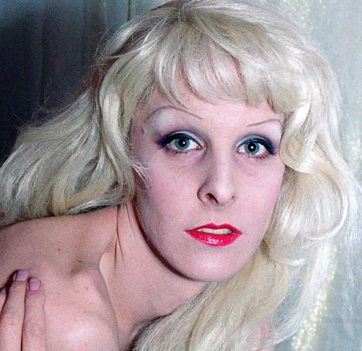 Greer Lankton photographed by Nan Goldin. She wears a long blonde wig, black winged eyeliner, light blue eyeshadow, and coral lipstick.