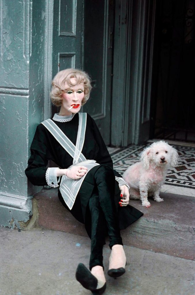 Sissy doll perched on the step of Einsteins. She wears a blonde pin-curled wig and a French maid style outfit.