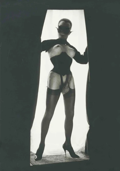 Le Chaman, a self-portrait photographed by Pierre Molinier circa 1967. A masked Molinier stands between two curtains. He is depicted with breasts and an erection. 