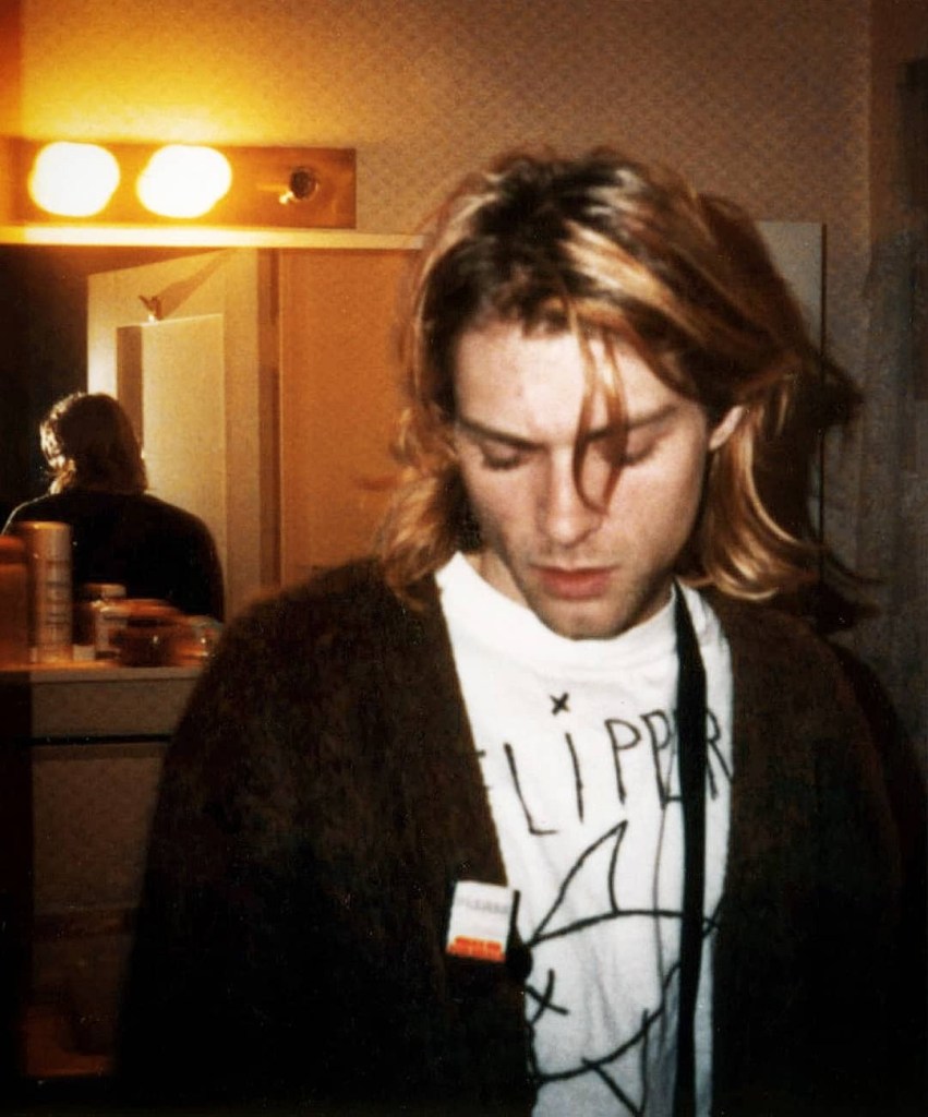 Kurt Cobain photographed by his wife, Courtney Love, in 1992. He's looking down and wears a brown cardigan and Flipper t-shirt.
